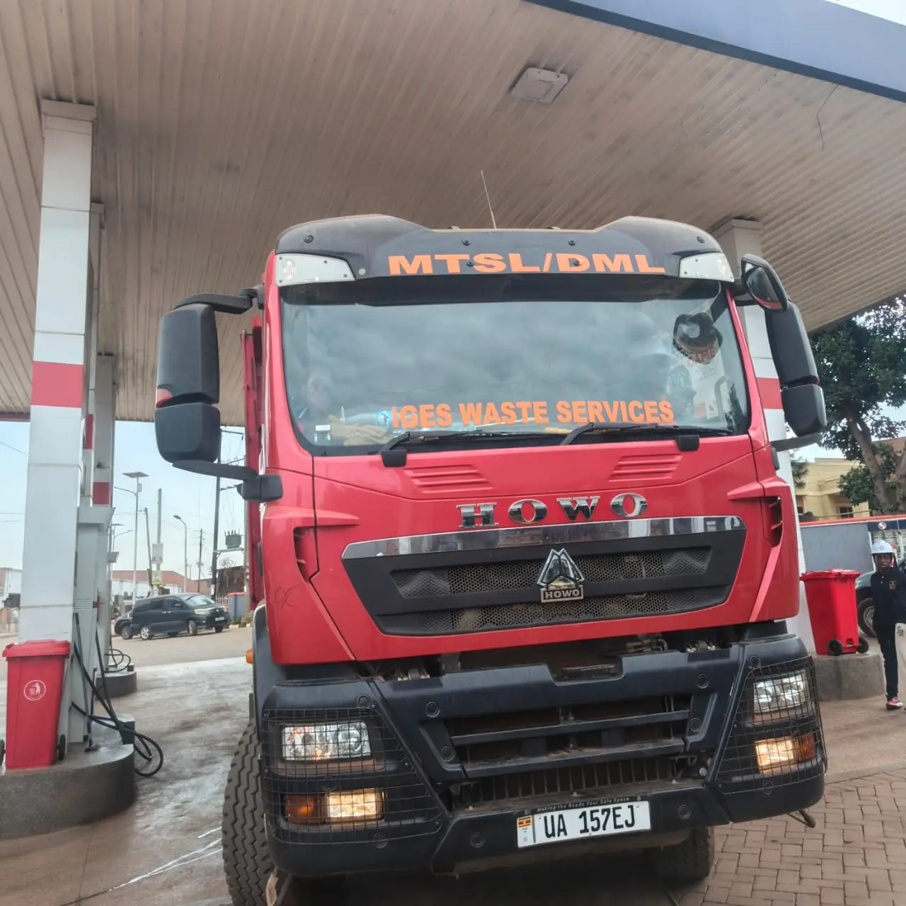 The front of a red HOWO truck lettered MTSL/DML and IGES WASTE SERVICES at a fuel station