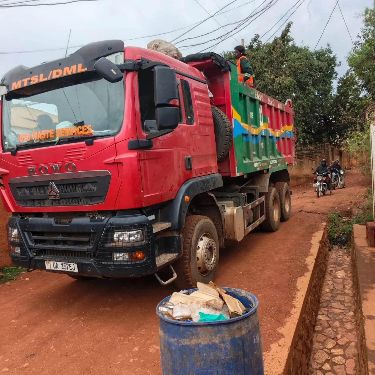 A red HOWO tipper lettered MTSL/DML in a narrow residential lane beside a blue bin