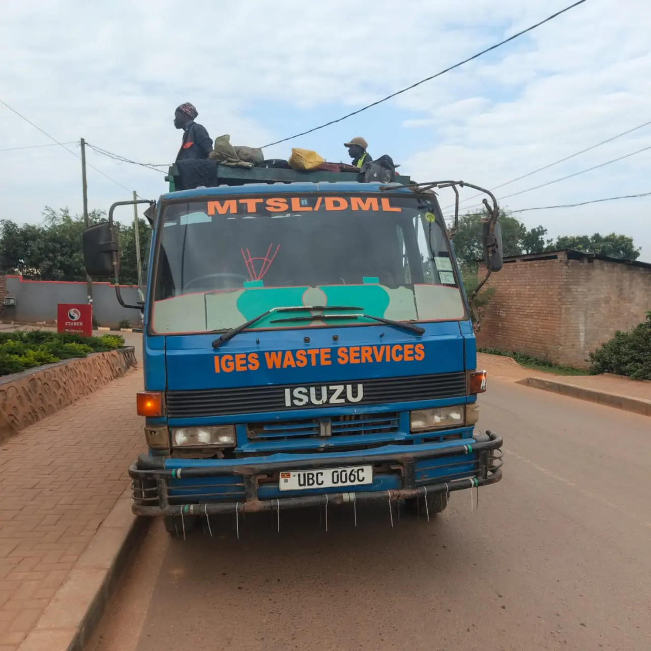 A blue Isuzu collection truck lettered IGES WASTE SERVICES, with crew standing on its load